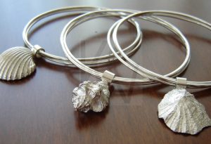 Brazalete artesanal de estilo semanario realizado en Plata de Ley 925, compuesto por tres aros independientes entrelazados que se mantienen unidos mediante un manguito; de este elemento de unión pende una concha marina detallada, pieza que varía según el diseño para ofrecer diferentes modelos de inspiración oceánica con un movimiento fluido y armónico.