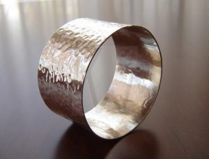 Brazalete artesanal de banda ancha realizado en Plata de Ley 925 con una superficie íntegramente martillada a mano; el acabado crea múltiples facetas que juegan con la luz para evocar los destellos y el movimiento natural del agua en un río.