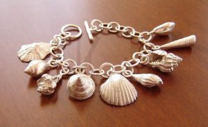 Pulsera artesanal compuesta por una serie de aros de los que penden nueve colgantes en forma de concha realizados en plata masiva, cada uno con un diseño y relieve diferente; el diseño captura la esencia marina mediante el movimiento de sus elementos, todos fabricados en Plata de Ley 925 con un relieve detallado.