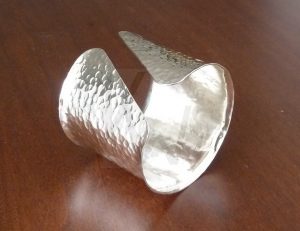 Brazalete artesanal de banda ancha en Plata de Ley 925 con superficie martillada y un distintivo corte diagonal en sus extremos; el diseño presenta los laterales ligeramente elevados, creando un relieve que enmarca los destellos de luz inspirados en la corriente del agua.