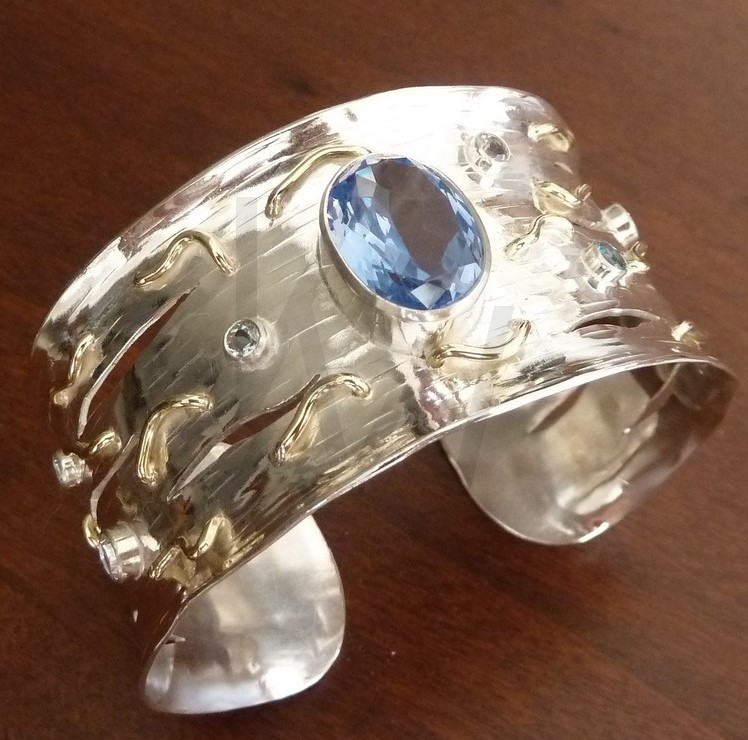 Brazalete de banda ancha en plata martillada e hilos de oro con una gran gema central azul y piedras naturales brillantes, evocando el movimiento de las olas y el brillo del mar.