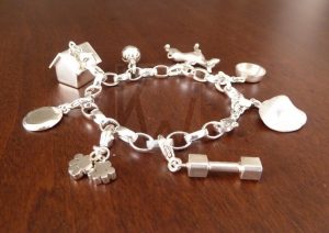Pulsera artesanal de plata diseñada con una cadena de eslabones que evoca la estética de un collar canino; de la estructura penden ocho colgantes detallados: una caseta, un cuenco, un cono de entrenamiento, un apport, una pelota, dos huellas de patas, un hueso, la silueta de un perro y una trufa, todo realizado en Plata de Ley 925.