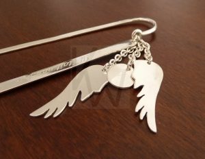 Marcapágina artesanal de Plata de Ley 925 con un diseño que presenta tres cadenas cortas suspendidas de un asa exterior; dos cadenas portan colgantes en forma de ala y la tercera sostiene un disco personalizable, mostrado con el número 7, pero disponible para grabar otras cifras, iniciales o símbolos que custodian el libro desde su lomo.