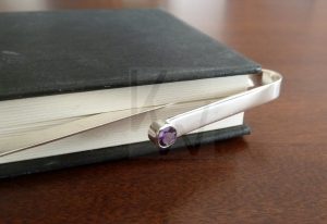 Marcapágina artesanal realizado en Plata de Ley 925 que presenta una gema natural engastada en el extremo que sobresale del libro; el diseño mostrado incluye una amatista, pero permite la personalización con otras piedras como granate, citrina, peridoto, topacio o cuarzo, aportando un toque de color y distinción sobre el lomo mientras custodia el punto de lectura.