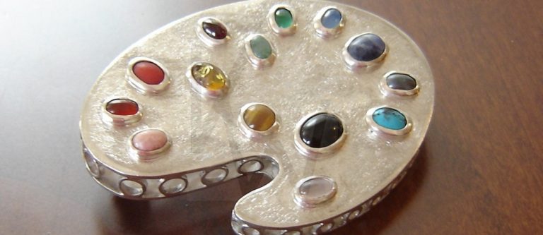 Broche artesanal de plata en forma de paleta de pintor con galería y gemas en talla cabujón de diversos colores, representando la creatividad y el espíritu artístico del joyero.