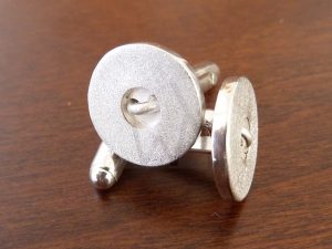 Gemelos artesanales realizados en Plata de Ley 925 que recrean con realismo la forma de un botón clásico; el diseño presenta un acabado satinado grueso y muestra un detalle de hilo de plata atravesando los dos orificios centrales, como si estuviera cosido al puño de la camisa en un elegante guiño a la sastrería tradicional.