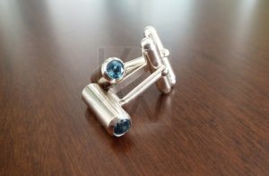 Pareja de gemelos artesanales realizados en Plata de Ley 925 con terminación pulida de alto brillo; la parte decorativa presenta un diseño en forma de tubo cilíndrico que alberga un topacio azul de 5mm engastado en cada extremo. Esta pieza de alta camisería permite la personalización de las gemas, ofreciendo opciones como granate, peridoto, citrino, amatista o cuarzo para adaptarse al estilo de cada ocasión.