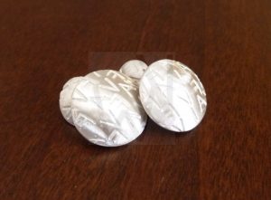 Pareja de gemelos artesanales realizados en Plata de Ley 925 con acabado mate satinado; cada pieza consta de dos discos convexos de distinto tamaño unidos por una fina cadena, donde el disco mayor presenta iniciales estampadas en toda su superficie, creando un juego de relieves y texturas personalizadas.