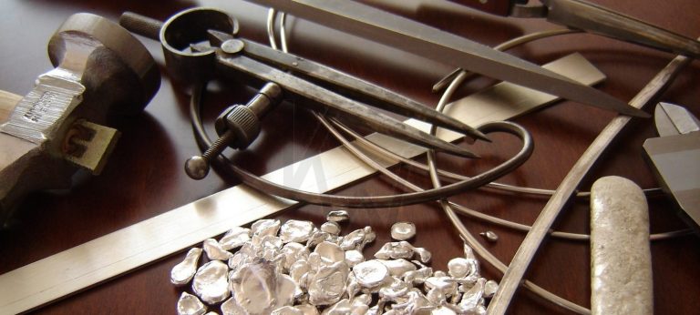 Detalle de materiales de joyería como plata en chapa, hilo y pepitas, junto a herramientas de precisión como compás, calibre y martillo, ilustrando los componentes del proceso creativo.