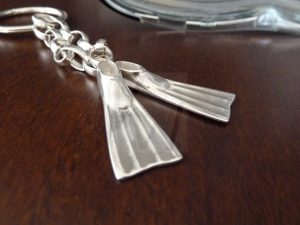 Llavero artesanal de Plata de Ley 925 compuesto por un aro principal del que parten dos cadenas de diferentes longitudes; cada cadena sostiene en su extremo una aleta de buceo detallada, creando un diseño dinámico y deportivo que rinde homenaje al mundo del submarinismo con la solidez de la plata masiva.
