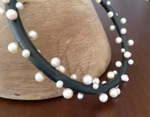 Collar artesanal compuesto por una base de caucho negro de acabado mate con cierre de Plata de Ley 925; sobre el cordón se distribuyen múltiples perlas blancas de diversos tamaños que crean un ritmo visual asimétrico y elegante.