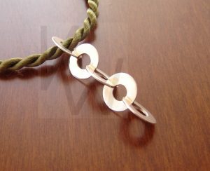 Colgante artesanal de plata compuesto por una estructura vertical de cinco discos fijos, dispuestos cada uno a 90 grados respecto al anterior para crear un efecto tridimensional, presentado sobre un cordón de seda trenzado en color verde oliva.