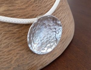 Colgante artesanal de plata con forma de disco ligeramente cóncavo y superficie martillada que evoca el movimiento del agua, presentado sobre una cadena de Plata de Ley 925 de diseño trenzado.