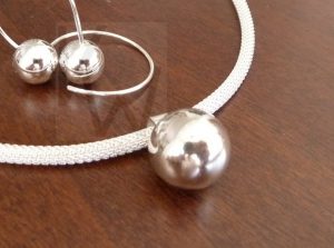Colgante artesanal de plata con forma de esfera sólida y pulida, presentado sobre una cadena de Plata de Ley 925 de diseño trenzado; al fondo de la imagen se muestra un par de pendientes a juego que completan el conjunto.