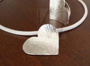 Colgante artesanal de plata con forma de corazón y superficie con textura martillada, presentado sobre una cadena de Plata de Ley 925 de diseño trenzado; al fondo de la imagen se aprecia un anillo a juego con el mismo acabado artesanal.