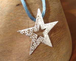 Colgante artesanal de plata con forma de estrella principal sobre la cual se encuentran estampadas múltiples estrellas de menor tamaño, creando un relieve detallado, presentado sobre un cordón azul que evoca el cielo nocturno.