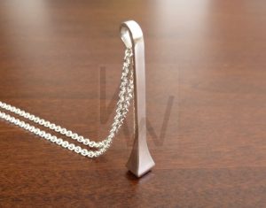 Colgante artesanal de plata con la forma de un clavo de herradero estilizado, de diseño alargado y acabado pulido, suspendido de una cadena de plata de eslabones finos que realza la elegancia de la pieza.