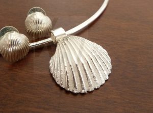 Conjunto de joyería artesanal en plata compuesto por un colgante en forma de concha sobre cadena rígida tipo omega, con pendientes a juego en segundo plano sobre fondo neutro.