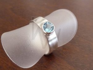 Anillo artesanal de plata con una banda ancha de 6 mm y acabado liso, que alberga un topacio azul de 5 mm engastado como punto central de luz y color.