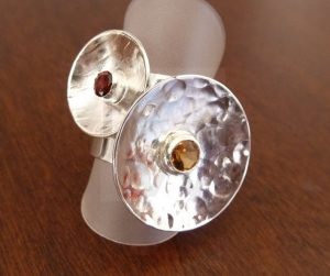 Pareja de anillos artesanales de plata con diseño circular cóncavo y superficie martillada, montados sobre bandas de 5 mm; uno de los anillos presenta un granate engastado en el centro y el otro un citrino, creando un contraste de reflejos y color.