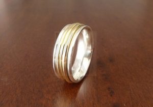 Anillo artesanal realizado en Plata de Ley 925 con una estructura de bordes elevados que actúan como rieles para tres aros independientes de Oro 18k; estos aros dorados giran libremente alrededor de la banda central de plata, creando un juego de movimiento, sonido sutil y contraste cromático que simboliza una unión dinámica y eterna.