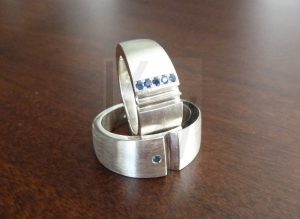 Pareja de dos alianzas artesanales de plata que, al unirse, completan la figura de un corazón dividido entre ambas piezas; la pieza con la parte superior del corazón presenta una fila de cinco pequeños zafiros engastados, mientras que la pieza con la parte inferior cuenta con un único zafiro incrustado, creando un conjunto de joyas coordinado y elegante.